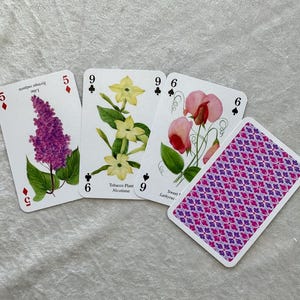 May include: A deck of playing cards with floral illustrations. Three cards show botanical drawings of lilac, tobacco plant, and sweet pea. The fourth card displays a purple and pink geometric pattern. The cards are on a light gray surface.