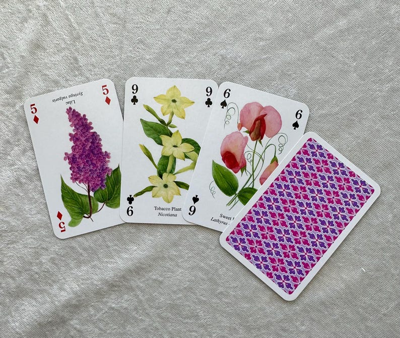 May include: A deck of playing cards with floral illustrations. Three cards show botanical drawings of lilac, tobacco plant, and sweet pea. The fourth card displays a purple and pink geometric pattern. The cards are on a light gray surface.