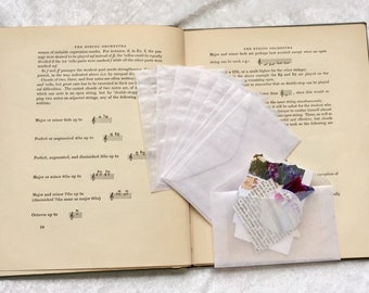 May include: An open book with a white page showing musical notation and a stack of white envelopes with a small bundle of colorful paper scraps on top of the book.