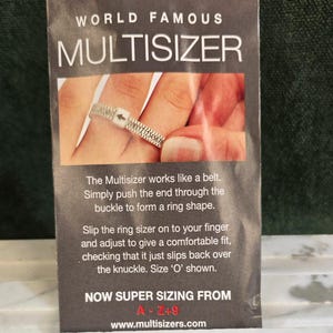 Op de afbeelding: Afbeelding van een Multisizer ringmaat. Het product is wit met zwarte tekst en is om een vinger gewikkeld. De tekst op het product luidt "WORLD FAMOUS MULTISIZER" en "NOW SUPER SIZING FROM A-Z+9".
