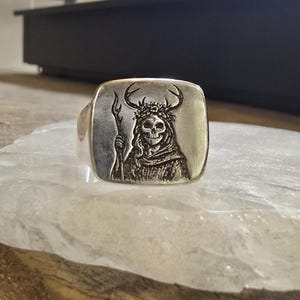 May include: A silver ring with a square face featuring a detailed engraving of a skeletal figure wearing a crown of leaves and antlers, holding a staff. The ring has a polished finish and a wide band.