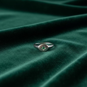 May include: A silver signet ring with a starburst design and a small, green gemstone at the center. The ring is set against a dark green, velvet-like fabric, creating a contrast that highlights the jewelry.
