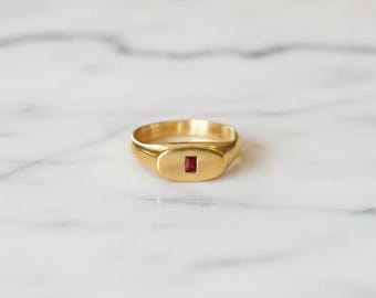 May include: A gold signet ring featuring a rectangular, deep red gemstone. The ring has a smooth, polished finish and a classic, minimalist design. The gemstone is set in the centre of the ring's face. The ring is shown on a white marble surface.