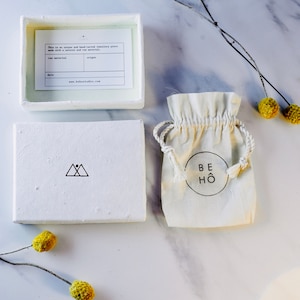 以下が含まれることがあります： 三角形のロゴと「This is an unique and handcrafted jewellery piece made with a natural and raw material.」というテキストが書かれた白い箱。箱が開いており、中には「BEH&Ouml;」というテキストが書かれた白い巾着袋が入っています。