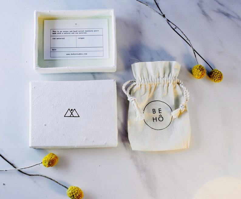 以下が含まれることがあります： 三角形のロゴと「This is an unique and handcrafted jewellery piece made with a natural and raw material.」というテキストが書かれた白い箱。箱が開いており、中には「BEH&Ouml;」というテキストが書かれた白い巾着袋が入っています。