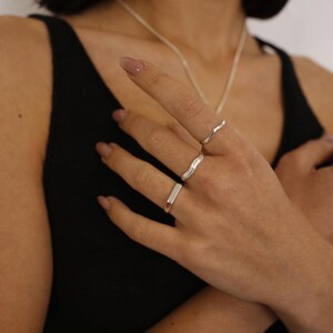 May include: A hand wearing three silver rings. The rings are stacked on the middle finger. The top ring is a thin, wavy band. The middle ring is a thicker, wavy band. The bottom ring is a straight, rectangular band.
