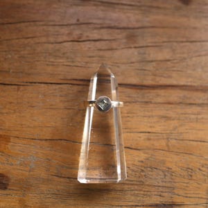 May include: A silver ring featuring a square, pale green gemstone within a circular setting. The ring is presented on a clear crystal point, set against a wooden backdrop. The ring's band is slender and plain.