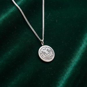 May include: A silver necklace with a circular pendant. The pendant features a detailed engraved design of hands, waves, and stars. The chain is a delicate curb style, and the pendant is centered. The background is a dark green velvet.
