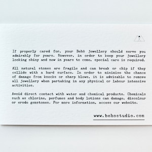 以下が含まれることがあります： ジュエリーのお手入れ方法を説明した白いカードに黒い文字が書かれています。テキストには、天然石の損傷を防ぐ方法や、ジュエリーを水や化学薬品から保護する方法が記載されています。カードには、ウェブサイトのアドレス www.behostudio.com も記載されています。
