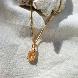 May include: A gold-toned chain necklace featuring a single oval-shaped, translucent orange gemstone pendant held in a four-prong setting. The pendant hangs from a delicate link chain, showcasing a bright, reflective metallic finish throughout.