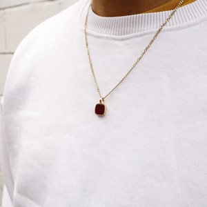 Puede incluir: Un collar de cadena dorada con un colgante cuadrado de color rojo oscuro. El colgante está engastado en un marco dorado. El collar se lleva sobre una sudadera blanca de cuello redondo.
