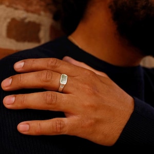 May include: A silver ring with a rectangular, light green stone is worn on a finger. The ring has a small star design on the side. The person is wearing a dark blue sweater. The background is a brick wall.