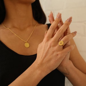 May include: A gold ring with a star design on a round face. The ring is worn on the index finger of a person's hand.