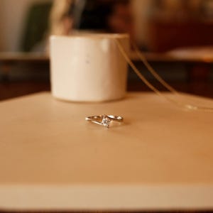 Könnte beinhalten: Ein silberfarbener Ring mit einem einzelnen, kleinen, quadratisch geschliffenen, klaren Stein in einer Ringschiene im Bypass-Stil. Die Schiene schwingt sich um den zentralen Stein und erzeugt ein fließendes, durchgehendes Design auf einer glatten Fläche.