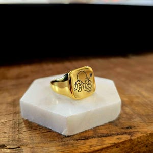 May include: A gold-colored signet ring with a square face. The face of the ring features a black octopus illustration. The ring is displayed on a white, hexagonal surface, set against a wooden background.