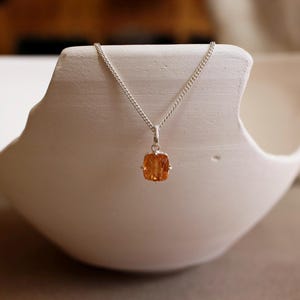 May include: A silver chain necklace featuring a rectangular, faceted orange gemstone pendant. The stone is set in a simple silver prong setting, suspended from a delicate, polished silver link chain that rests against a smooth, curved white surface.