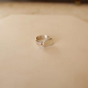 Puede incluir: Un anillo de sello plateado con una cara rectangular y una pequeña gema roja. El anillo se encuentra sobre un fondo neutro, resaltando su diseño simple y elegante. Una pieza de joyería clásica.