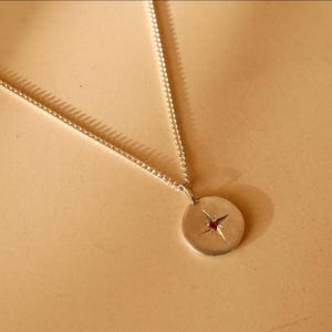 May include: A silver-coloured metal necklace featuring a circular pendant with a four-pointed star engraving at the centre. A small, dark red gemstone is set in the middle of the star. The pendant hangs from a delicate, thin silver-coloured chain.