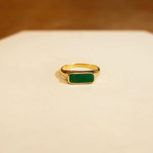 May include: A gold ring featuring a rectangular, emerald-green stone. The ring has a classic design with a polished finish. The band is a standard width, and the stone is set flush with the ring's surface. A stylish piece of jewellery.