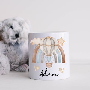 May include: White ceramic mug featuring a watercolour illustration of a hot air balloon soaring over a rainbow with stars. The name "Adam" is written beneath the balloon.