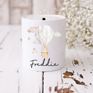 May include: White ceramic money box with a hot air balloon design. The balloon is grey and white with a brown basket. There are two small animals in the basket. The background is white with brown clouds and gold stars. The name Freddie is written in black below the balloon.