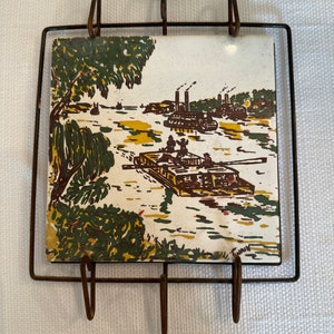 Puede incluir: Un azulejo vintage con una escena pintada de barcos en un río, enmarcado en un estante de metal con ganchos. El azulejo es blanco con acentos verdes, marrones y amarillos.