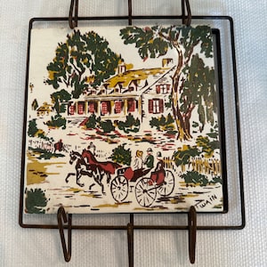 Puede incluir: Un azulejo de cerámica vintage con una ilustración colorida de una casa, árboles y un carruaje tirado por caballos. El azulejo está enmarcado en un bastidor de metal con tres ganchos.