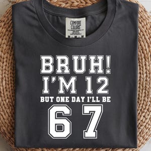 Könnte beinhalten: Dunkelgraues T-Shirt mit dem Text "BRUH! I'M 12 BUT ONE DAY I'LL BE 67" in weißen Blockbuchstaben. Das T-Shirt ist gefaltet und liegt auf einem gewebten, kreisförmigen Platzset vor hellem Holzhintergrund.
