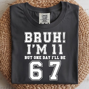 Könnte beinhalten: Dunkelgraues T-Shirt mit dem weißen Text "BRUH! I'M 11 BUT ONE DAY I'LL BE 67". Das Shirt liegt gefaltet auf einer runden Matte aus Korbgeflecht vor hellem Holzgrund.