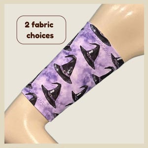 May include: A fabric arm sleeve featuring a repeating pattern of black witch hats on a mottled purple background. The text 2 fabric choices is displayed in a brown outlined box in the upper left corner of the image.