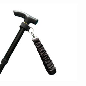 Puede incluir: Bastón negro con un mango en forma de T y una correa para la muñeca. El bastón tiene un eje negro con un anillo dorado. La correa para la muñeca está hecha de tela negra fruncida y se sujeta con un cierre plateado.