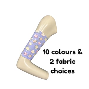 Puede incluir: Un brazo de maniquí beige con una funda de tela de color morado claro que presenta un patrón repetitivo de pequeñas tortugas amarillas y estrellas rosas. El texto 10 colours & 2 fabric choices aparece impreso en negro junto al brazo.