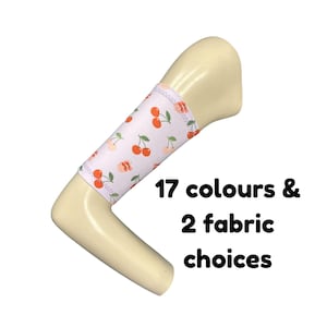 Puede incluir: Una funda para mango en forma de L de color crema con una cubierta de tela blanca que presenta un patrón repetitivo de cerezas rojas y hojas verdes. El texto 17 colours & 2 fabric choices está impreso en negro junto a la funda.