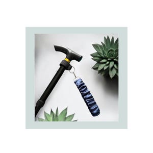 Puede incluir: Bastón de marcha negro ajustable con un mango en forma de T y una correa de muñeca azul marino. El bastón tiene un acento dorado cerca del mango. La correa de muñeca es de un material fruncido y sedoso. Sobre fondo blanco con suculentas verdes.