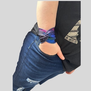 May include: A galaxy print wristband with a blue, pink, and purple color scheme. The wristband is tied in a knot and worn on a person's wrist.
