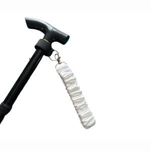 Puede incluir: Un bastón negro ajustable con un mango en forma de T y una correa de muñeca de tela blanca fruncida, sujeta con un cierre plateado. El bastón está diseñado para ayudar a la movilidad.