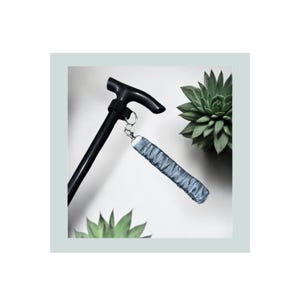 Puede incluir: Un bastón negro con un mango curvo, con una correa de muñeca gris sujeta por una cadena plateada. La correa está hecha de una tela texturizada. El fondo incluye plantas suculentas verdes sobre un fondo blanco.