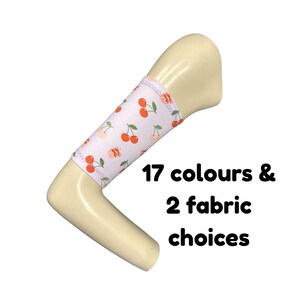 Puede incluir: Una funda para mango en forma de L de color crema con una cubierta de tela blanca que presenta un patrón repetitivo de cerezas rojas y hojas verdes. El texto 17 colours & 2 fabric choices está impreso en negro junto a la funda.