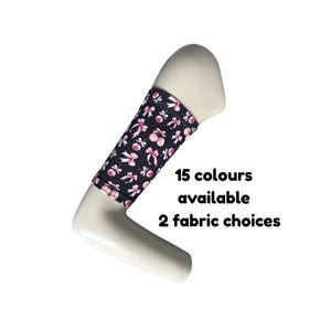 Puede incluir: Una manga para brazo de color negro con un patrón repetitivo de lazos y cerezas rosas. El texto 15 colours available 2 fabric choices está impreso junto a la manga. La manga se muestra sobre un maniquí de brazo de color beige.