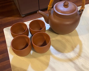 May include: A brown ceramic tea set with a teapot and four matching cups. The teapot has a bamboo handle and a lid with a small knob. The cups are round and open. The set is on a light-colored surface.