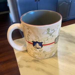 May include: A beige ceramic mug with a white handle and a green interior. The mug features a design with Mickey Mouse, a shield, and the text "2000 Disneyland". A red stripe runs across the mug.