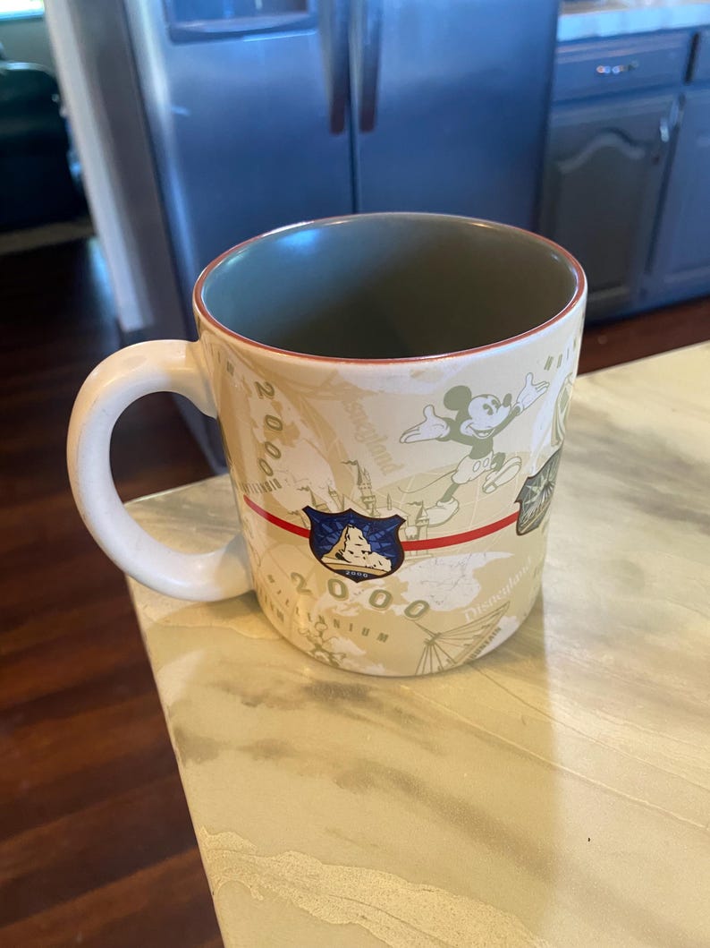 May include: A beige ceramic mug with a white handle and a green interior. The mug features a design with Mickey Mouse, a shield, and the text "2000 Disneyland". A red stripe runs across the mug.