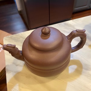 May include: A brown ceramic teapot with a rounded body, a lid with a small knob, and a curved handle. The spout and handle ends are sculpted with dragon head details. The teapot is sitting on a light-colored surface.