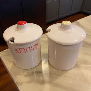 May include: Two white ceramic condiment jars with red and yellow lids. The jar on the left says "KETCHUP" in red lettering. The jar on the right says "MUSTARD" in yellow lettering.