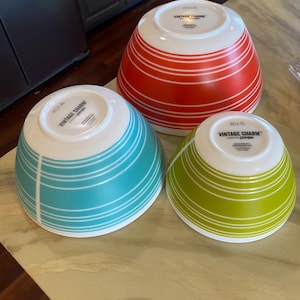 May include: Three Pyrex Vintage Charm mixing bowls in red, teal, and green, each with white horizontal stripes. The bowls are upside down, revealing the white interiors and the "VINTAGE CHARM" logo.