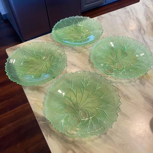 May include: A set of four light green glass plates, each featuring a detailed embossed leaf pattern with a textured, serrated edge. The plates are translucent and circular, showcasing a prominent central vein design that radiates outward to the rim.