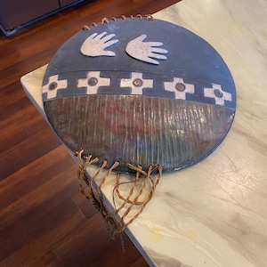 May include: A circular wall hanging featuring a dark gray upper section with two white handprints and a horizontal band of white cross symbols. The lower half has a textured, multi-colored finish with brown and green tones, accented by braided leather cords.