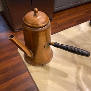May include: A cylindrical copper coffee pot with a matching lid featuring a small round knob. The pot has a straight, angled spout and a horizontal black handle with a ribbed texture. The copper surface shows a warm, metallic finish with patina.