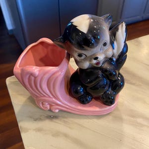 May include: A ceramic planter featuring a glossy black and white skunk figurine attached to a pink, shell-shaped vessel. The skunk has large eyes, a small nose, and a bushy tail, while the pink base displays decorative, swirling embossed details.