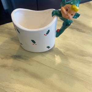 May include: A white ceramic mug featuring a scalloped rim and a pattern of small green holly leaves with red berries. The handle is shaped like a smiling elf wearing a green hat with a red tip and yellow hair, appearing to climb the side.